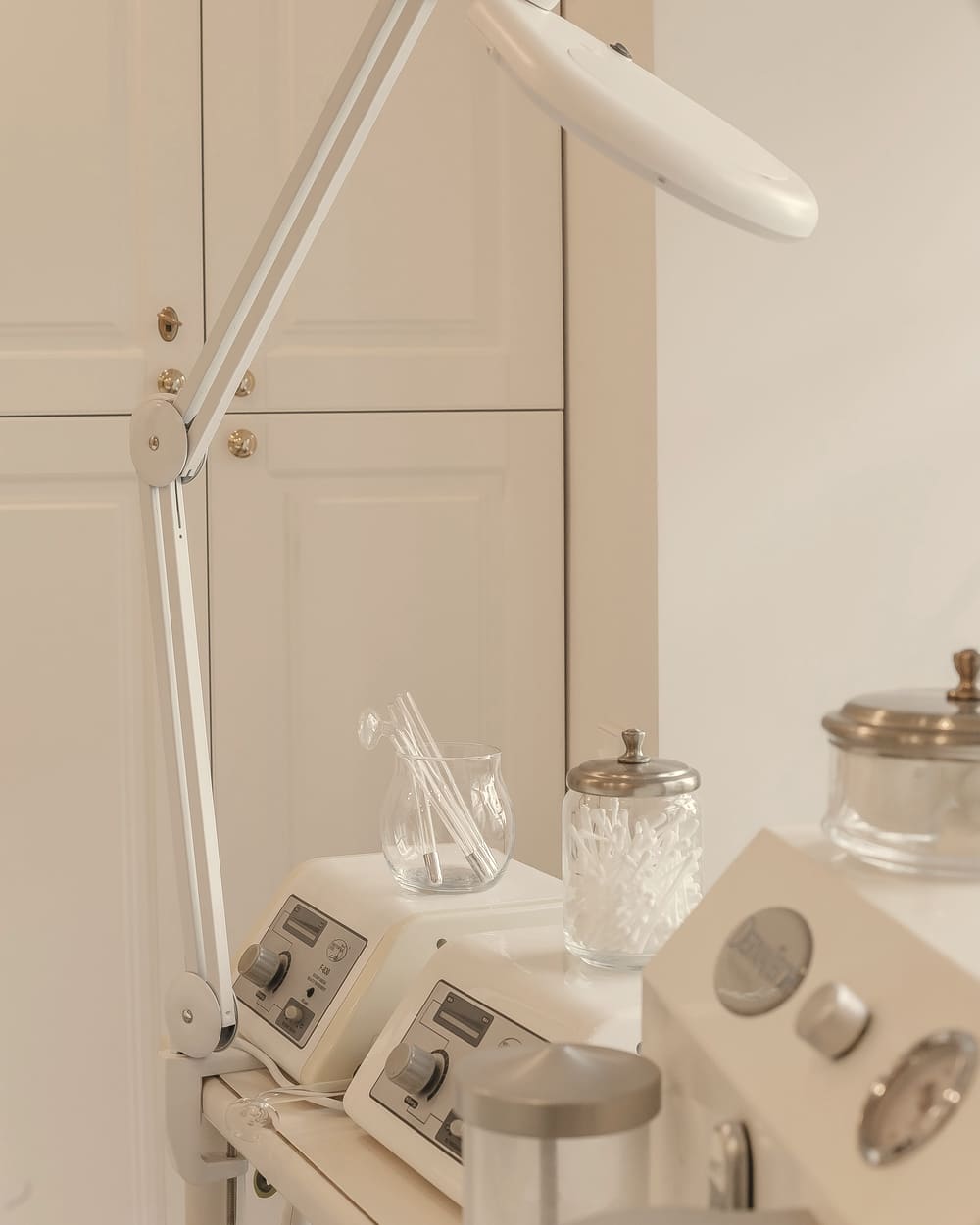 Cosmetology devices, a magnifying lamp and glass jars with metal lids beside light-coloured cabinets